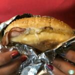 hands holding a bun with melted cheese and meat slices in aluminum foil