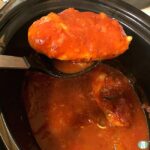 a piece of chicken lifted out of the slow cooker on a large spoon out of a red sauce