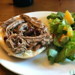 Pulled pork on an open face bun is placed on a white plate with a salad on the side, which contains chopped avocado, tomatoes and orange segments