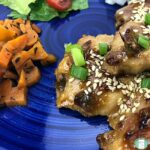 glazed chicken topped with green onions and sesame seeds next to carrots and a salad