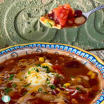 spoon lifting soup out of a bowl of red broth with corn, tomatoes, beans and topped with sour cream and cheese