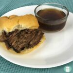 bun filled with beef on a plate next to a small bowl of brown au jus gravy