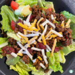 lettuce topped with diced tomato, diced avocado, kidney beans mixed with ground beef, and sprinkled with shredded cheese