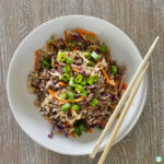 a meal with ground sausage, matchstick carrots and coleslaw and topped with chopped green onion is served in a white bowl with chopsticks sitting askew on the edge of the bowl