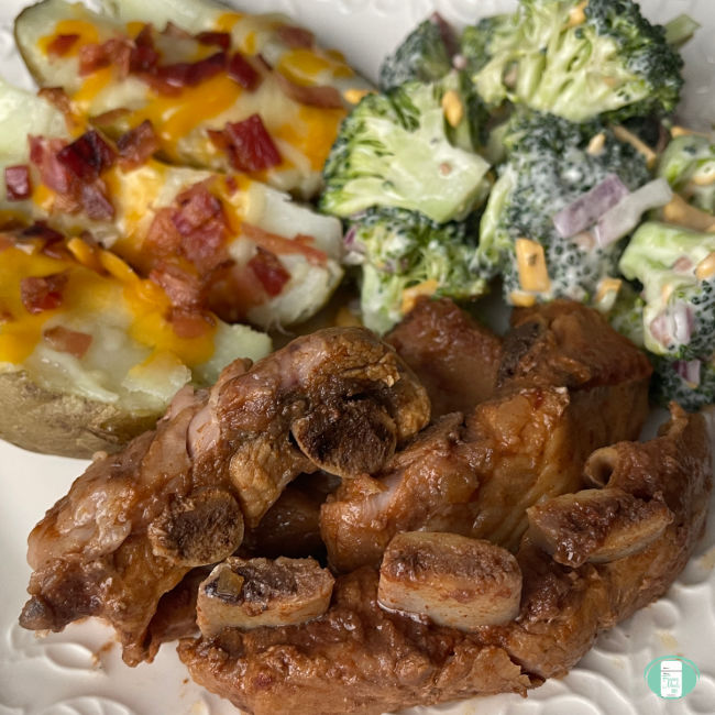 A few BBQ ribs on a plate, smothered in BBQ sauce. Served with a broccoli salad and potato skins covered with shredded cheese and crumbled bacon.
