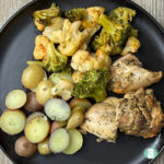 dark plate with halved baby potatoes, broccoli, cauliflower, and two pieces of chicken topped with herbs