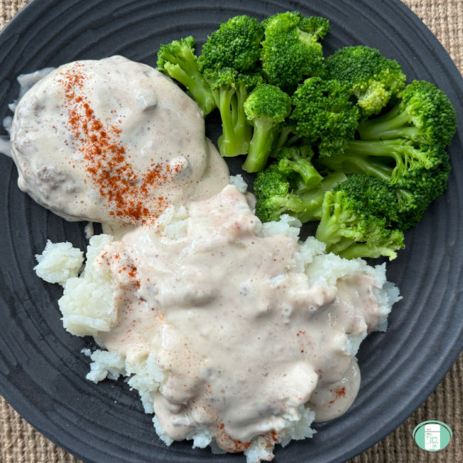 A beef patty and mashed potatoes smothered in a light coloured mushroom gravy and sprinkled with a splash of paprika, plated alongside steamed broccoli on a black plate.