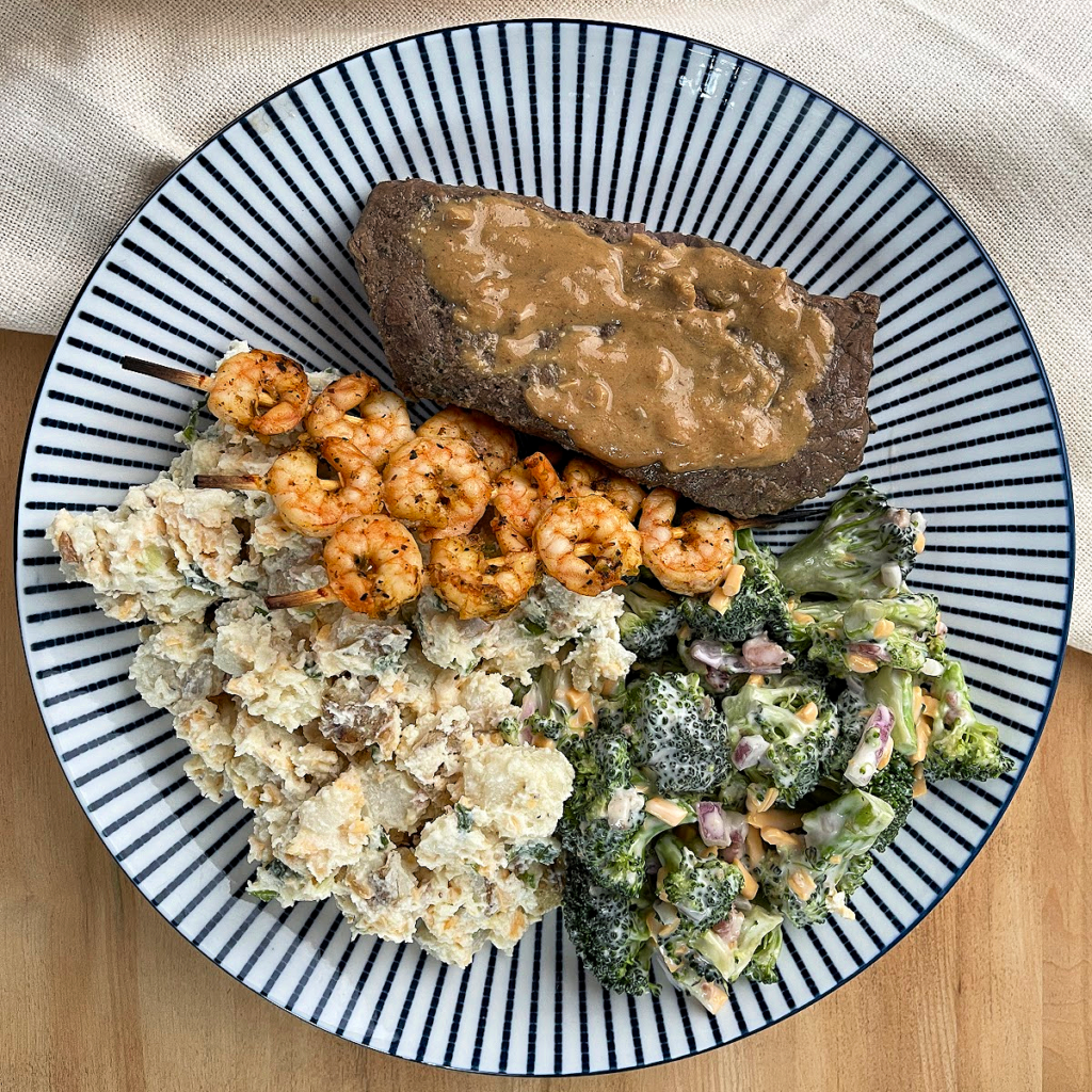 Steak smothered in sesame sauce, plated with skewers of cajun shrimp, potato salad and broccoli salad.