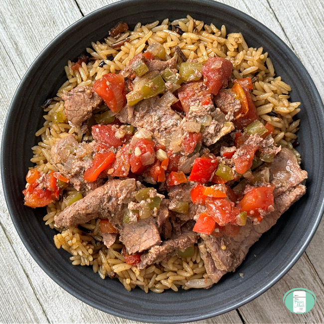 Beef strips served with diced tomatoes, red and green peppers on a bed of rice in a large black bowl.