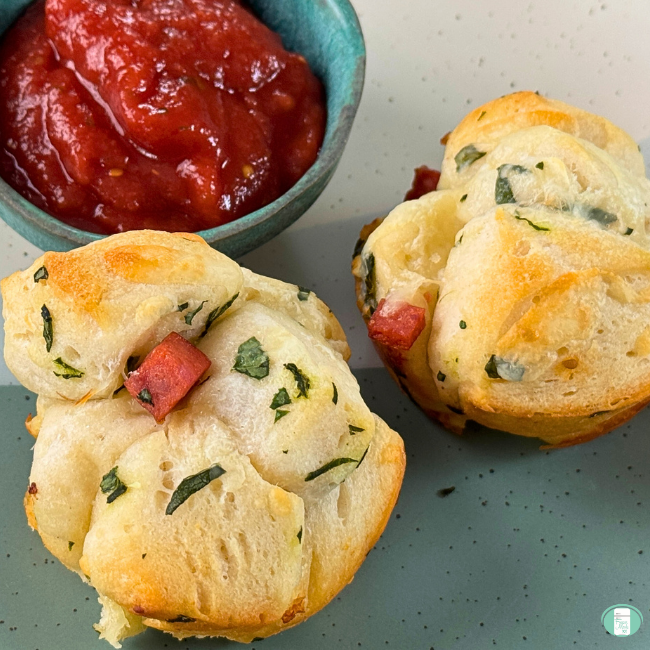 toasty little muffins with bits of pepperoni, basil and cheese served on a plate with a side of marinara sauce for dipping